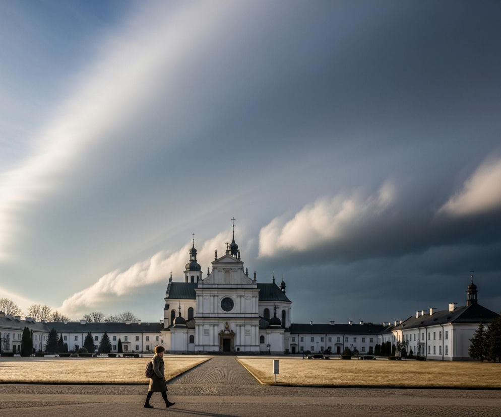 Kobieta z krótkimi rudymi włosami, ubrana w beżowy płaszcz i ciemne spodnie, idzie w prawą stronę brukowaną ścieżką. Przed nią rozciąga się duży, trawiasty plac o złotobrązowym odcieniu, otoczony z trzech stron białymi, klasycystycznymi budynkami z ciemnymi dachami. W tle dominuje jasna, dwuwieżowa bazylika z ciemnymi kopułami i zdobionym portalem, a za nią i po prawej stronie niebo pokrywają ciemne, wydłużone chmury, kontrastujące z jaśniejszym, błękitnym niebem po lewej stronie.
