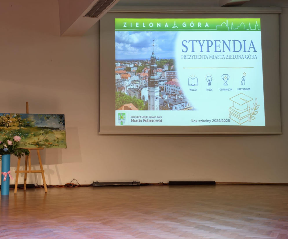 Uroczystość wręczenia miejskich stypendiów w auli uniwersytetu. Na ekranie prezentacja Stypendia Prezydenta Miasta Zielona Góra z panoramą miasta i ikonami symbolizującymi wiedzę, pasję, osiągnięcia oraz przyszłość. Obok stoi sztaluga z obrazem i wazon z kwiatami. Więcej o stypendiach przeczytasz na naszym portalu.