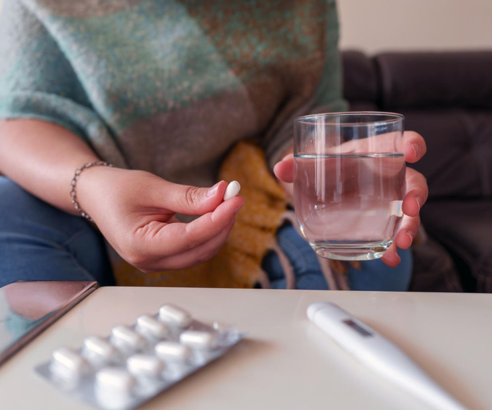 Kobieta trzymająca w dłoni białą tabletkę i szklankę wody, przygotowująca się do zażycia leku przeciwbólowego. Na stole opakowanie tabletek i termometr, co symbolizuje walkę z bólem. Ostrzeżenia o zagrożeniach związanych z nadmiernym zażywaniem tabletek na ból można przeczytać na portalu Poradnik Zdrowie.