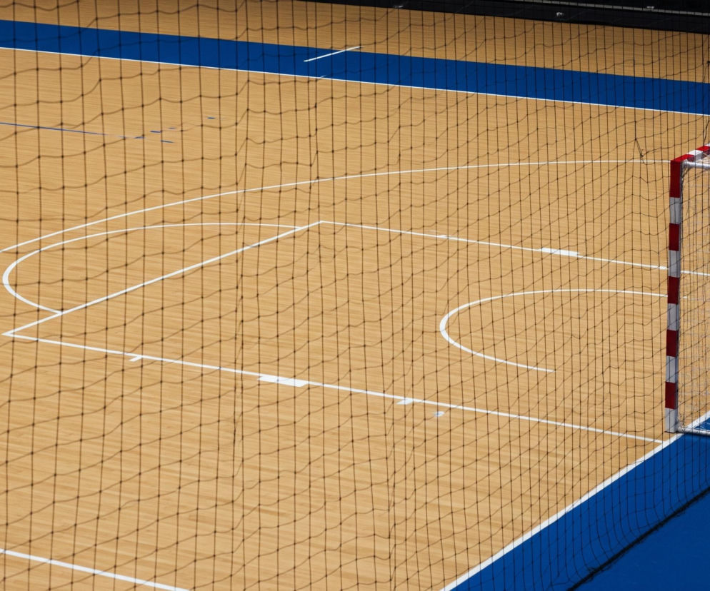 Centralny punkt zdjęcia zajmuje jasnożółte drewniane boisko sportowe z białymi liniami, które wyznaczają różne obszary gry, w tym łuki i linie proste. Na boisku, od góry do dołu, rozciąga się ciemna siatka, rzucająca regularne, romboidalne cienie. W prawym górnym rogu znajduje się bramka z biało-czerwonymi słupkami i poprzeczką, za którą rozciąga się biała siatka. Pod bramką i na krawędzi boiska widoczny jest szeroki pas intensywnie niebieskiej nawierzchni.