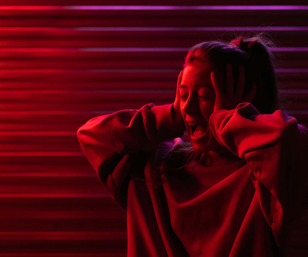 Młoda kobieta w czerwonej bluzie z długimi rękawami, trzymająca się za uszy i krzycząca z bólu lub irytacji, co symbolizuje problem szumów usznych. Tło stanowią poziome pasy w odcieniach czerwieni i fioletu. Dowiedz się więcej o przyczynach i leczeniu szumów usznych na portalu Poradnik Zdrowie.