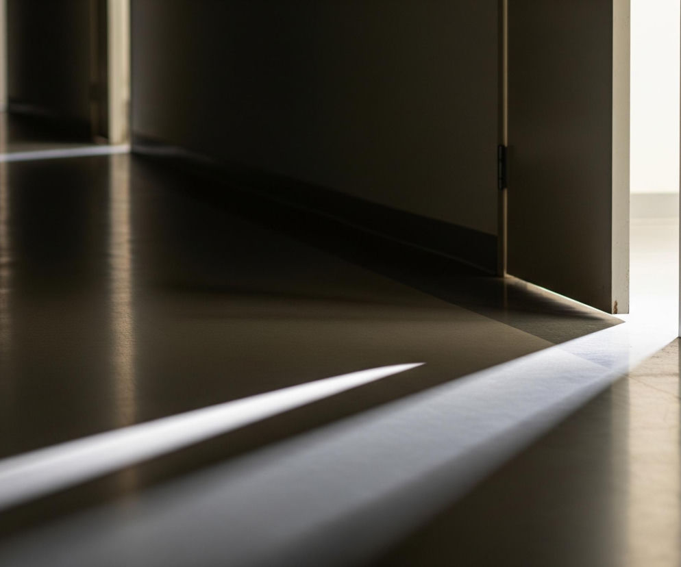 Jasne światło wydobywające się z lekko uchylonych białych drzwi po prawej stronie oświetla ciemny korytarz. Smuga światła biegnie od progu w głąb korytarza, tworząc na podłodze długi, trójkątny kształt, który zwęża się w oddali. Podłoga korytarza jest ciemna i lśniąca, odbijając blade refleksy światła. W oddali widać kolejne ciemne drzwi i fragmenty ścian.