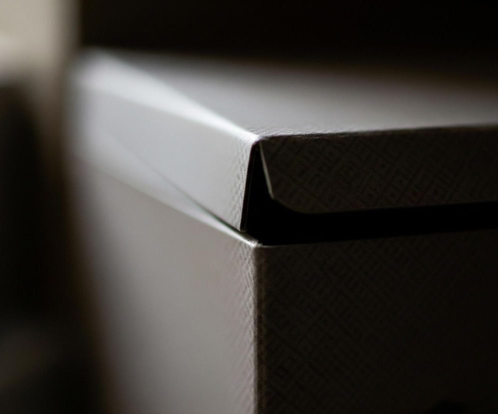 Szczegółowy plan przedstawia górny róg kartonowego pudełka o delikatnym, beżowo-szarym kolorze. Pudełko ma drobną, wytłaczaną teksturę tworzącą wzór małych rombów lub siatki. Górna pokrywa pudełka jest lekko odsunięta od jego głównej części, tworząc wąską, ciemną szczelinę. Światło pada z lewej strony, delikatnie oświetlając krawędź pokrywy i część boku, podczas gdy większa część obrazu pogrążona jest w cieniu, co nadaje kompozycji tajemniczy i stonowany charakter.