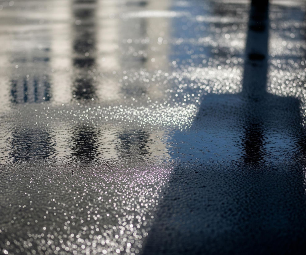 Powierzchnia asfaltu, pokryta licznymi drobnymi kropelkami wody, odbija światło, tworząc rozmyte bliki. W tle widoczne są odbicia zarysów budynku z oknami, rozmyte i nieostre. Po prawej stronie dominują cienie, a woda na asfalcie wydaje się ciemniejsza. Światło rozchodzi się po całej powierzchni, tworząc efekt bokeh na licznych, drobnych punkcikach.