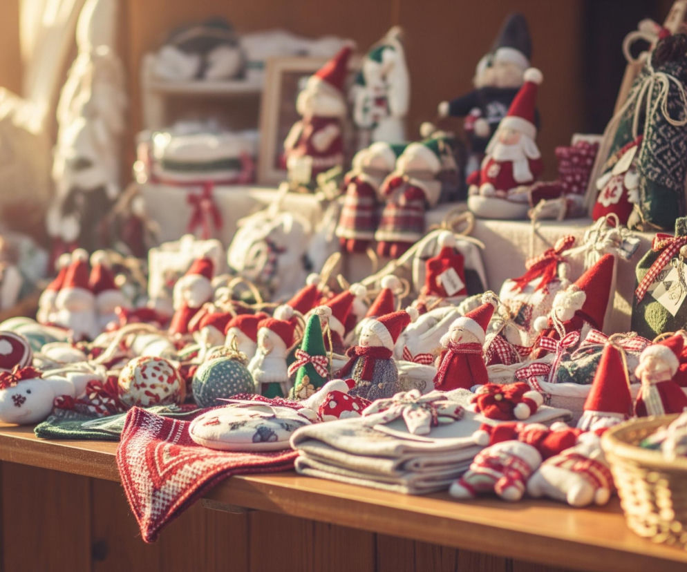 Drewniane stoisko jest wypełnione ręcznie robionymi ozdobami i upominkami, głównie w odcieniach czerwieni, bieli i zieleni. Na pierwszym planie leży złożona czerwono-biała tkanina z geometrycznym wzorem, obok niej stos beżowych, złożonych materiałów. Wśród dekoracji dominują figurki w kształcie krasnali i Mikołajów w czerwonych czapkach, a także różnorodne bombki i małe pakunki przewiązane wstążkami w czerwoną kratę. Całość jest skąpana w ciepłym, rozproszonym świetle słonecznym, tworzącym miękkie cienie i jasne refleksy.