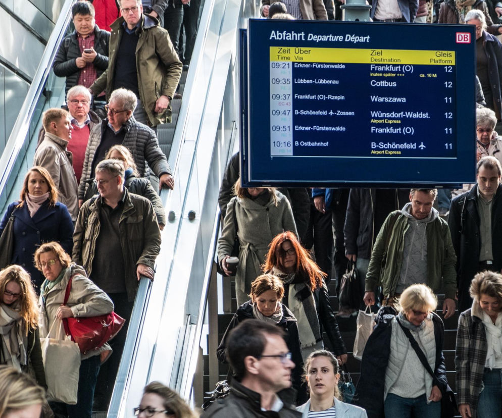 Ludzie z bagażami na ruchomych schodach na lotnisku, a w tle tablica odlotów wskazująca na utrudnienia w podróżowaniu. To ujęcie symbolizuje chaos i paraliż, o którym można przeczytać na portalu Super Biznes.