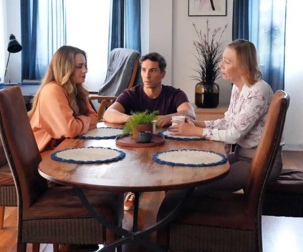 Oliwka (Wiktoria Gąsiewska) w pomarańczowej bluzie, Tomasz (Jakub Sokołowski) w fioletowej koszulce oraz Wanda (Maja Barełkowska) w białej bluzce z wzorami, siedzą przy drewnianym stole w pokoju z błękitnymi zasłonami, rozmawiając. Więcej o ich losach w Super Seriale.