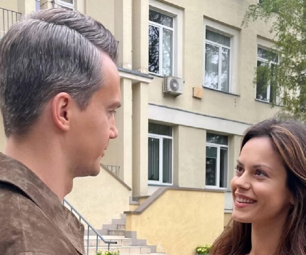 Paulina Lasota (Kasia) i Krzysztof Kwiatkowski (Jakub) uśmiechają się do siebie, stojąc na zewnątrz budynku. Kasia ma długie, brązowe włosy, a Jakub elegancko zaczesane, siwe. W tle widać fragment kremowej ściany budynku z oknami. Ten kadr zapowiada emocje w M jak miłość, o czym więcej na Super Seriale.