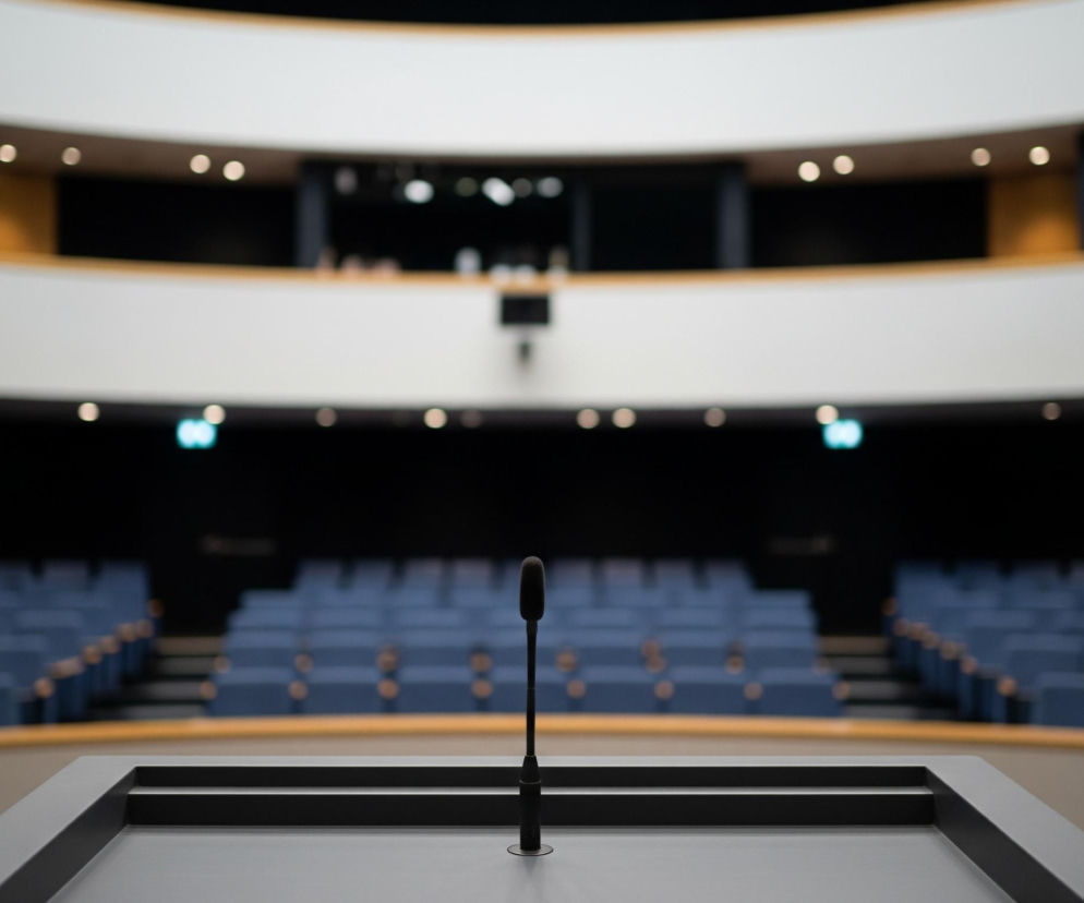 Czarny mikrofon na długiej, cienkiej nóżce stoi na szarym mównicy z czarnymi krawędziami. W tle rozciąga się puste audytorium z rzędami niebieskich foteli. Górna część pomieszczenia ukazuje dwa poziomy balkonów z kremowymi balustradami, podświetlonymi małymi, okrągłymi lampami.