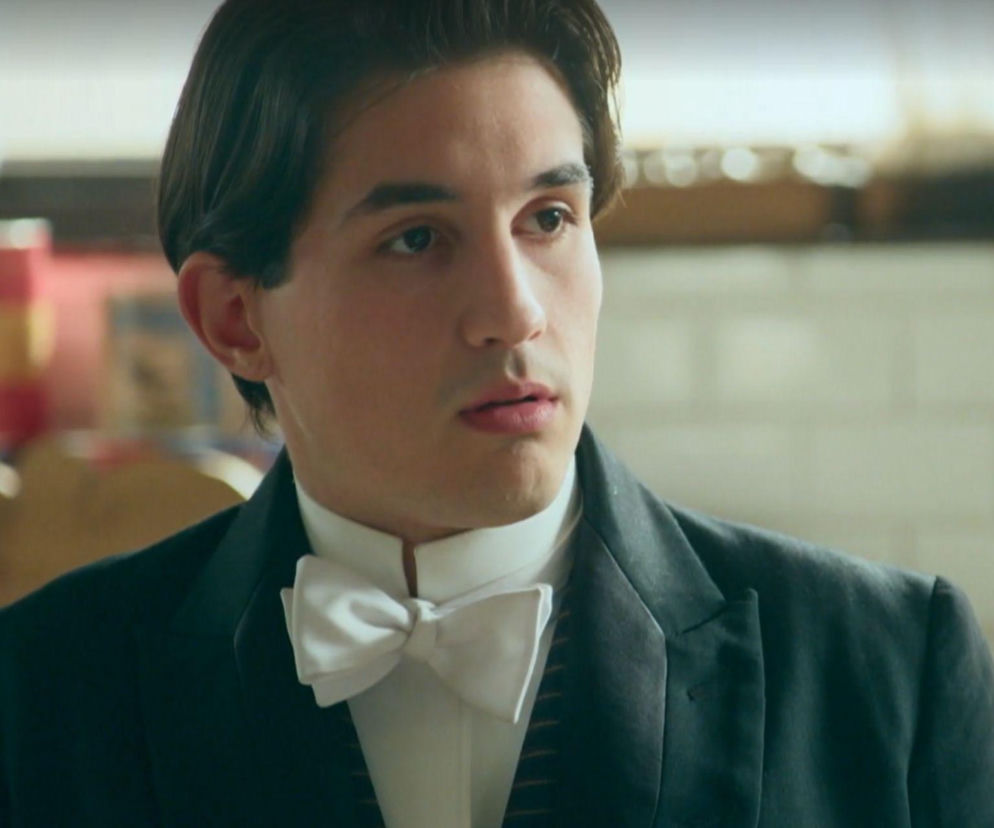 Młody mężczyzna, Curro, ubrany w elegancki czarny garnitur i białą muchę, o ciemnych włosach, spoglądający w prawą stronę z zamyśleniem. Postać z hiszpańskiego serialu La promesa, o której można przeczytać więcej na portalu Super Seriale.