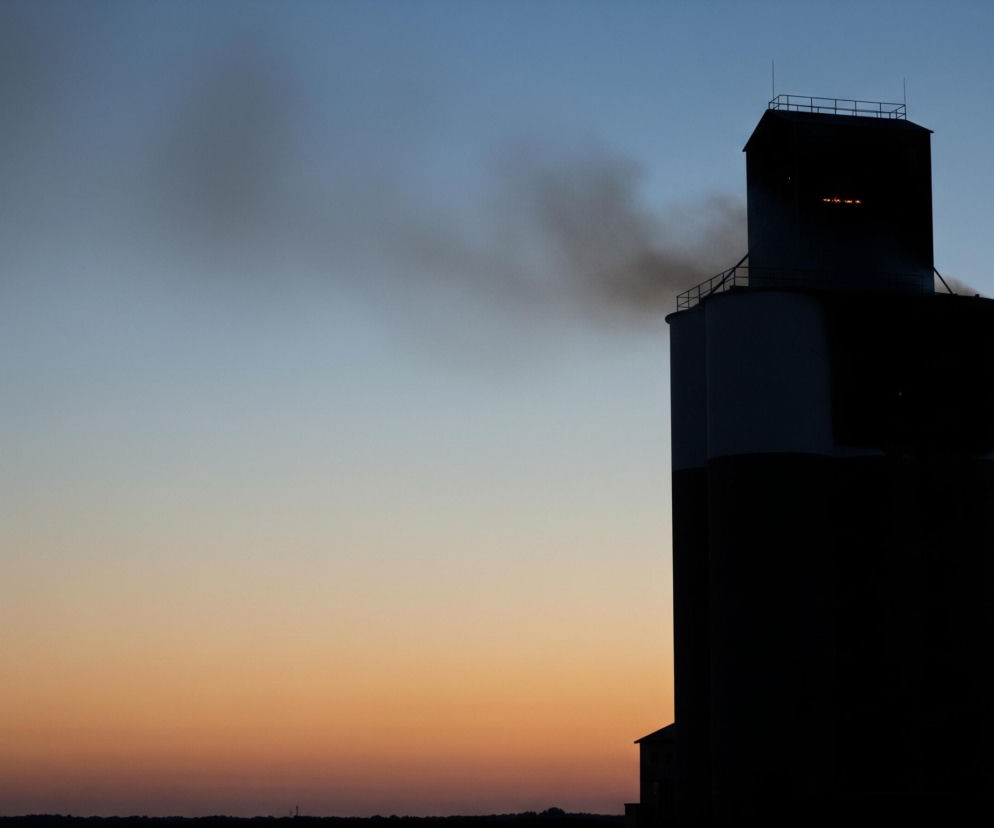Wysoki, ciemny silos zbożowy w prawym centrum obrazu dominuje nad horyzontem. Z lewej strony górnej części silosu wydobywa się ciemny dym, a w jego wnętrzu widać pomarańczowe światła. Tło stanowi stopniowo przechodzące niebo: od głębokiego błękitu u góry do jasnego, pomarańczowo-brązowego odcienia u dołu. Na dalekim horyzoncie widoczny jest ciemny zarys drzew i innych konstrukcji.