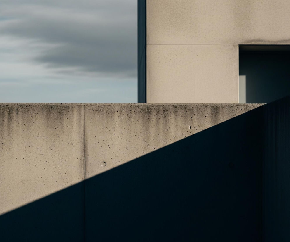 Architektoniczną kompozycję z elementami betonu i nieba. Na pierwszym planie znajduje się poziomy mur z szarego betonu, który przecina obraz na wysokości około jednej trzeciej od dołu. Na murze widać cieniowanie, gdzie lewa część jest oświetlona, a prawa pokryta głębokim, ukośnym cieniem, tworzącym geometryczny podział.

Powyżej muru, po prawej stronie, wznosi się fragment prostopadłościennej budowli z jasnoszarego betonu, w której znajduje się prostokątny, ciemny otwór przypominający wejście lub okno. Po lewej stronie, za murem, rozciąga się jasne, zachmurzone niebo. Geometria i kontrasty światła i cienia dominują w tej scenie.