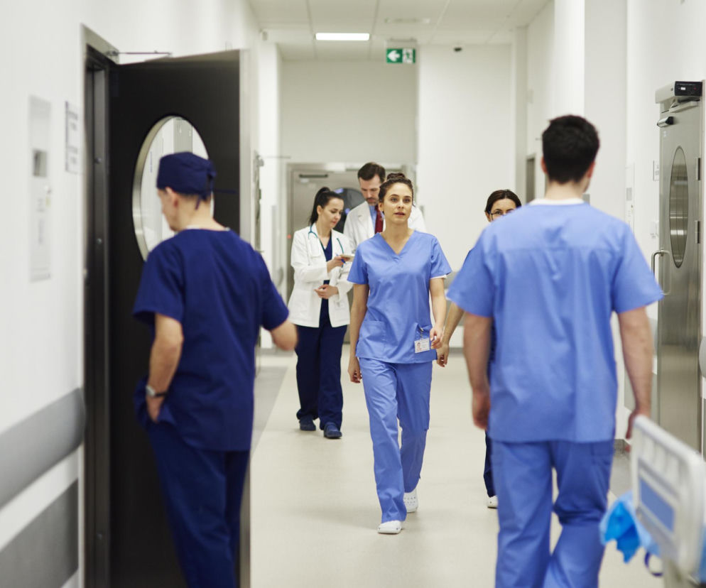 Personel medyczny w szpitalnym korytarzu, w tym pielęgniarki i lekarze, idący w różnych kierunkach, co symbolizuje codzienną pracę i wyzwania w placówkach zdrowia. Więcej o problemach szpitali powiatowych przeczytasz na Poradnik Zdrowie.