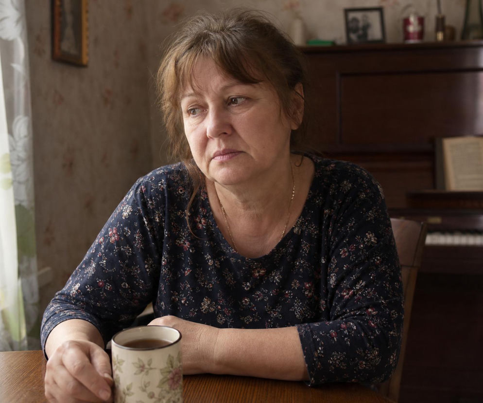 Zamyślona kobieta w średnim wieku, ubrana w granatową bluzkę w kwiaty, siedzi przy stole z kubkiem herbaty. W tle widać fragment pokoju z pianinem. Cała historia jej rozterek jest dostępna na naszym portalu.