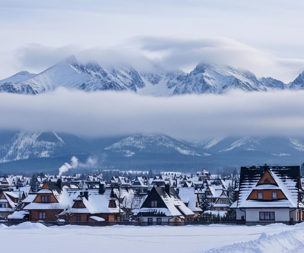 Zasypane śniegiem szczyty górskie, częściowo przesłonięte białymi chmurami, dominują na horyzoncie pod jasnoszarym niebem. Poniżej rozciąga się pasmo niższych, ciemniejszych gór, a następnie rozległa dolina pokryta grubą warstwą śniegu. W centrum znajduje się skupisko tradycyjnych drewnianych domów z ciemnymi dachami, również obficie pokrytych śniegiem, z których niektórych kominów wydobywa się dym. Na pierwszym planie, po prawej stronie, widać kilka osób idących zaśnieżoną ścieżką, a po lewej rozciąga się zaśnieżone pole.