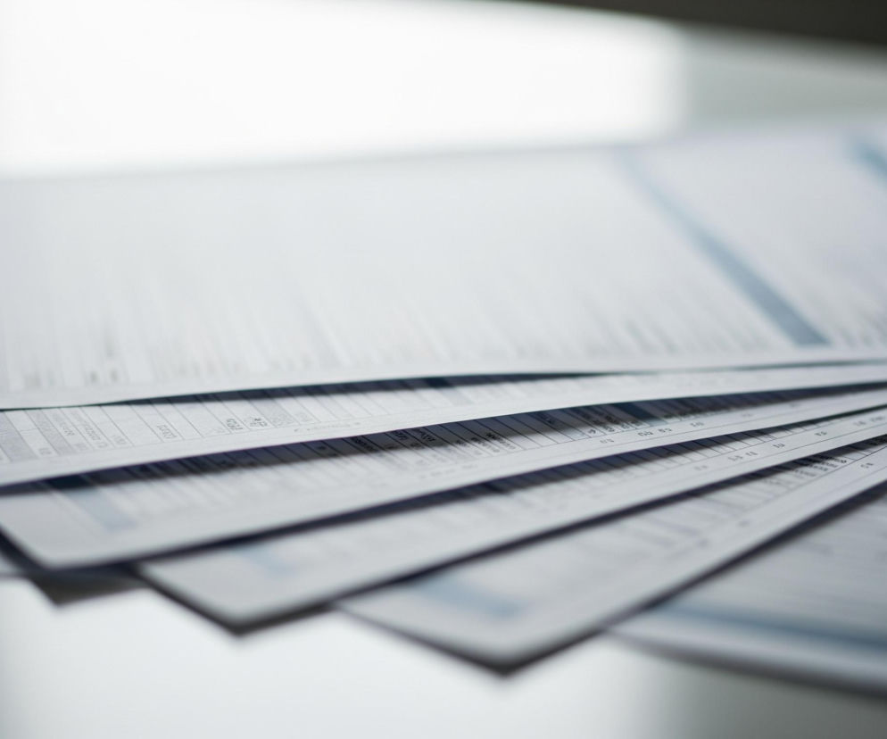 Stos papieru w białych i jasnoszarych odcieniach, leżący na jasnej powierzchni, zajmuje większość kadru. Kartki są rozłożone wachlarzowo, z widocznymi poziomymi liniami i kolumnami tekstu lub danych na każdej z nich. Na górze stosu kartki są lekko rozmazane, a w tle widać rozmyte, jasne światło.