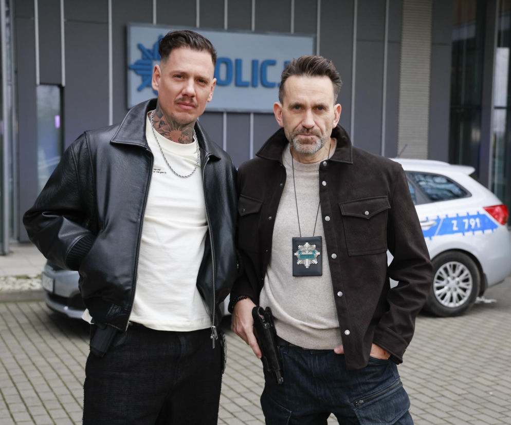 Aktorzy Aleksander Mackiewicz jako Adam Bogusz i Radosław Rutkowski jako Krzysztof Chrobociński, ubrani w cywilne ubrania, pozują przed budynkiem policji. W tle widać radiowóz. Więcej o ich przygodach w serialu Gliniarze znajdziesz na Super Seriale.