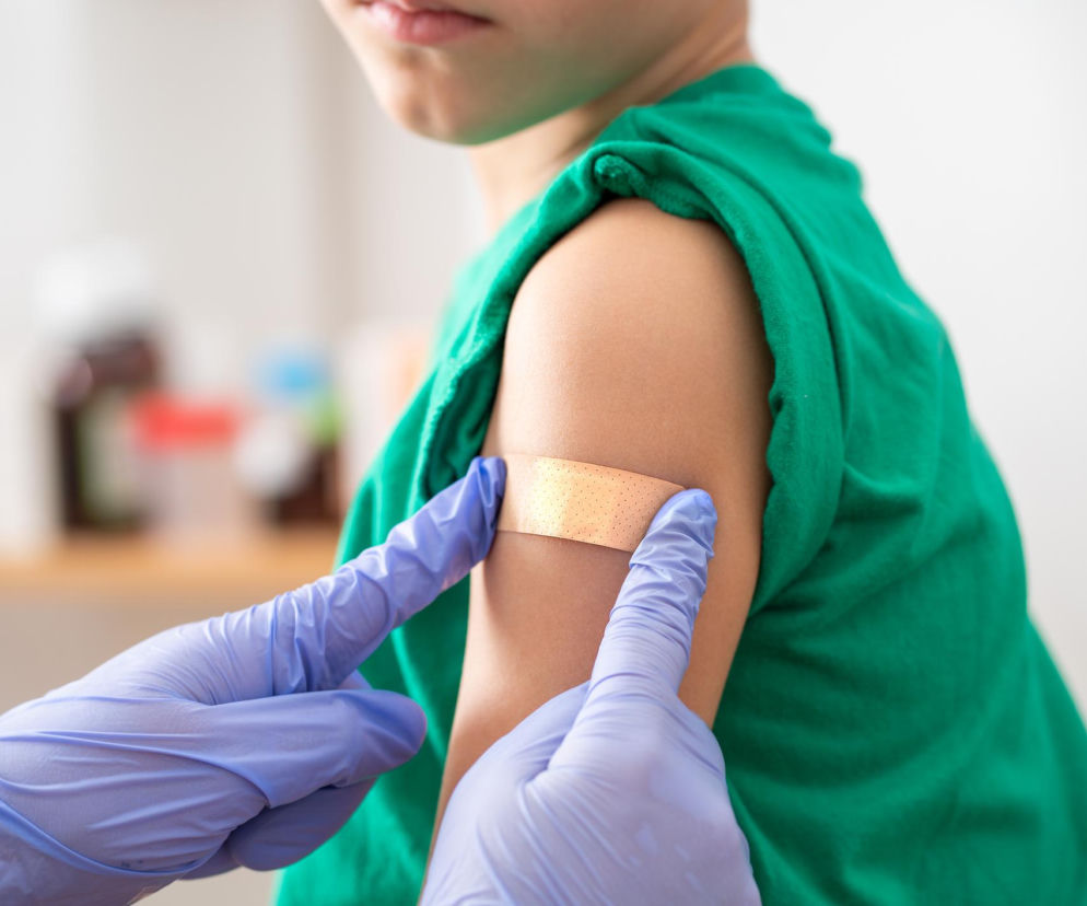 Dziecko w zielonej koszulce ma plaster na ramieniu po szczepieniu. Ręce w fioletowych rękawiczkach medycznych delikatnie dotykają plastra. Poradnik Zdrowie informuje o nowym systemie namierzania dzieci bez szczepionek.
