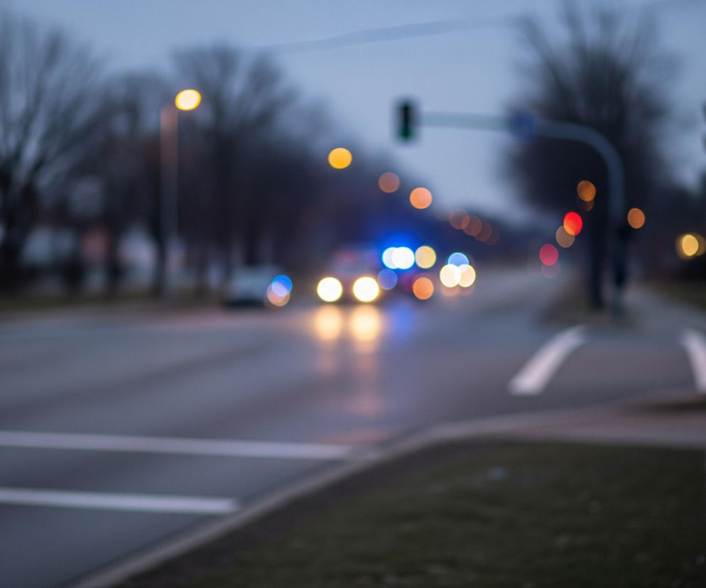 Rozmazana scena uliczna przedstawia drogę asfaltową z przejeżdżającymi samochodami i oświetleniem ulicznym o zmierzchu. Po lewej stronie widoczne są światła tylne samochodów i czerwone światło sygnalizacji, a w oddali niebieskie, migające światła sugerują pojazd uprzywilejowany. Prawa strona kadru ukazuje chodnik i latarnię uliczną, z niewyraźnymi zarysami budynków w tle. Całość jest skąpana w ciemnych, chłodnych barwach, z ciepłymi punktami świetlnymi.