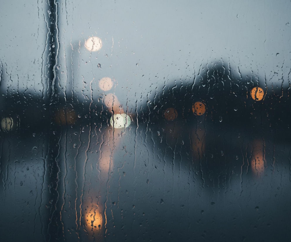 Szybę pokrytą gęsto kroplami deszczu i spływającymi smugami wody, tworzącymi dynamiczne wzory. Tło jest rozmyte, zdominowane przez szarość i ciemnoniebieskie odcienie, z wyraźnymi, rozświetlonymi okrągłymi plamami światła w kolorze białym, żółtym i pomarańczowym. Pionowy, ciemny element, być może słupek lub ramka, jest widoczny po lewej stronie, przecinając obraz od góry do dołu. Całość tworzy wrażenie patrzenia przez mokrą szybę na nocny lub pochmurny krajobraz miejski.