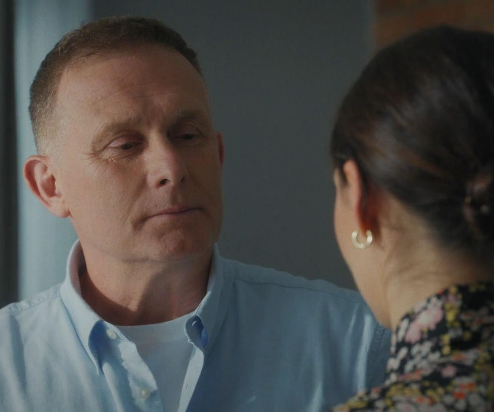 Mężczyzna w jasnoniebieskiej koszuli, Artur Rogowski, patrzy na Joannę Dobrzańską, ubraną w kwiecistą bluzkę, tyłem do widza. Scena z serialu M jak miłość przedstawiająca Artura i Joannę, którzy spędzają razem czas, co jest kluczowe w fabule Super Seriale.