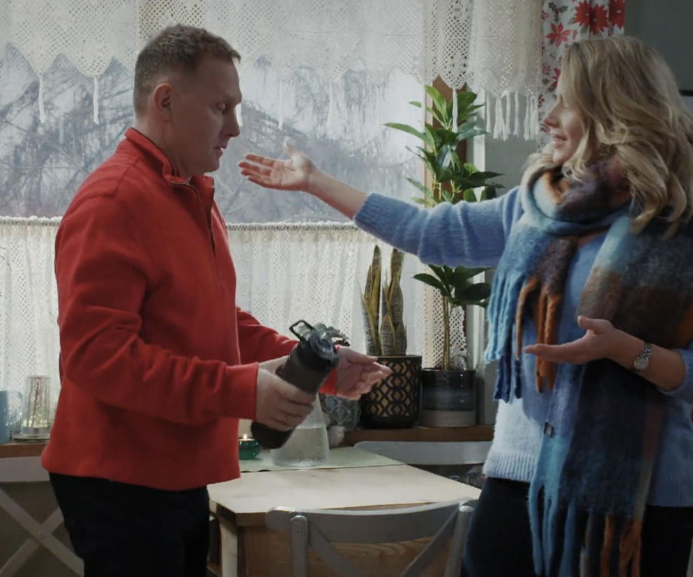 Artur Rogowski (Robert Moskwa) w czerwonej bluzie trzymający bidon i Judyta (Paulina Chruściel) w niebieskim swetrze oraz szalu, rozmawiający w kuchni o zmianach w życiu Artura, co opisują Super Seriale.