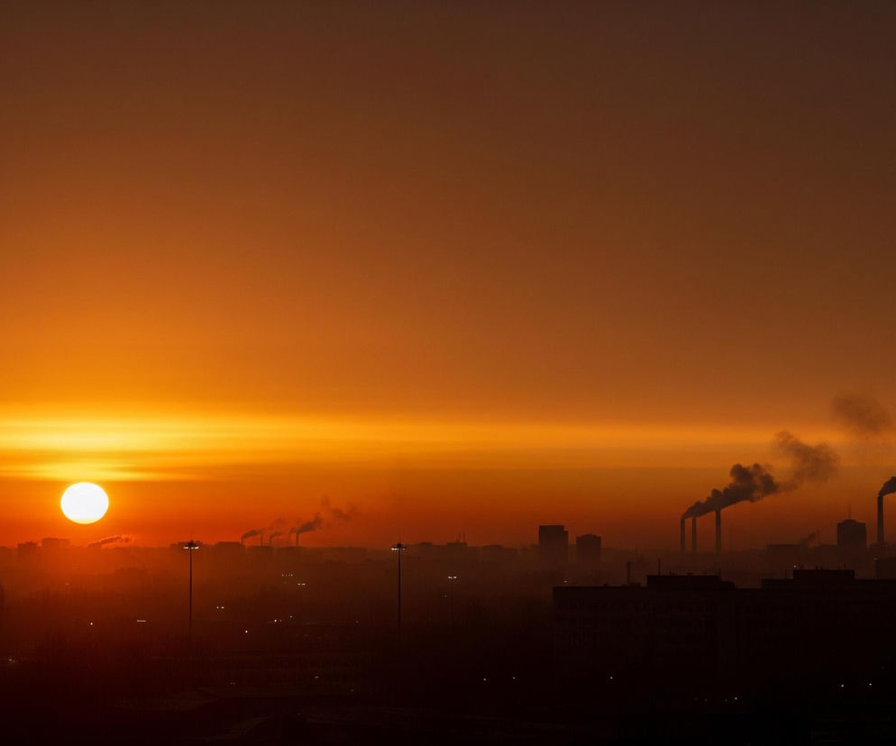 W centrum kadru, na tle pomarańczowego nieba, widoczne jest jasno świecące, białe słońce, które tworzy wyraźną poziomą smugę światła na horyzoncie. Poniżej rozciąga się ciemna sylwetka miejskiego krajobrazu, z licznymi budynkami i kominami fabrycznymi, z których unosi się dym w prawym górnym rogu. Cała scena jest zdominowana przez ciepłe odcienie pomarańczu i brązu, przechodzące od jasnego blasku słońca do ciemniejszych barw u góry i u dołu obrazu.