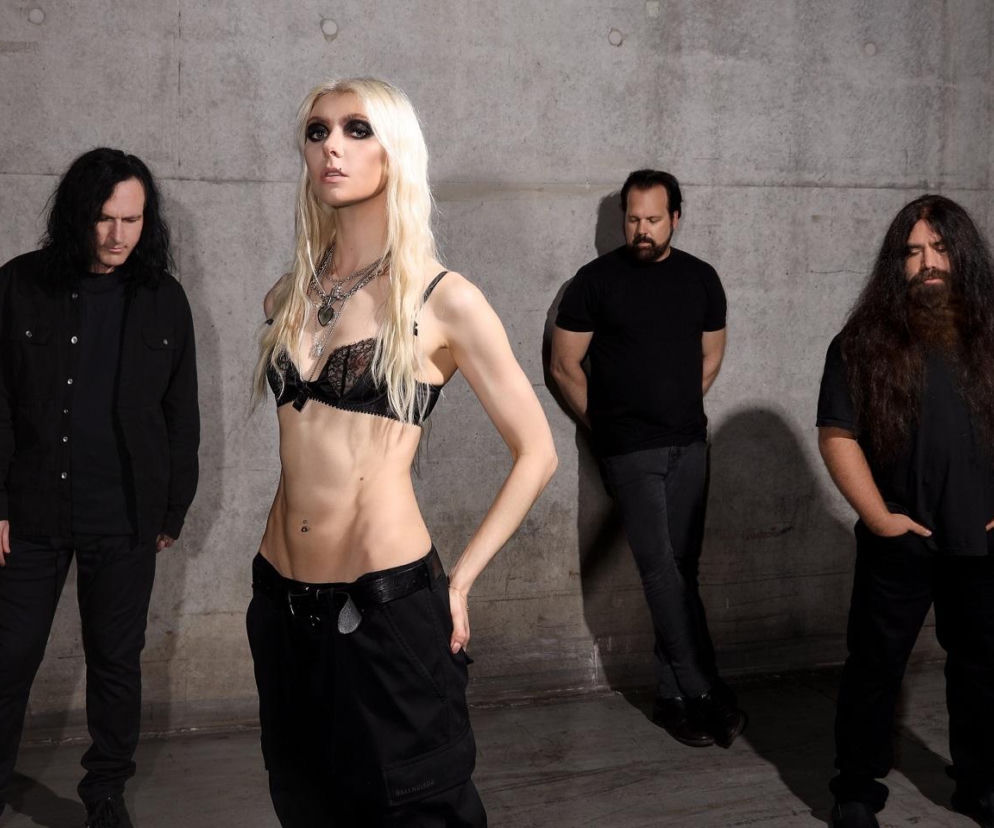 Zespół The Pretty Reckless, od lewej: Mark Damon, Taylor Momsen, Ben Phillips i Jamie Perkins, pozuje na tle betonowej ściany. Taylor, blondynka w czarnym biustonoszu i szerokich spodniach, stoi przodem. Więcej o ich nowym albumie Dear God przeczytasz na naszym portalu.
