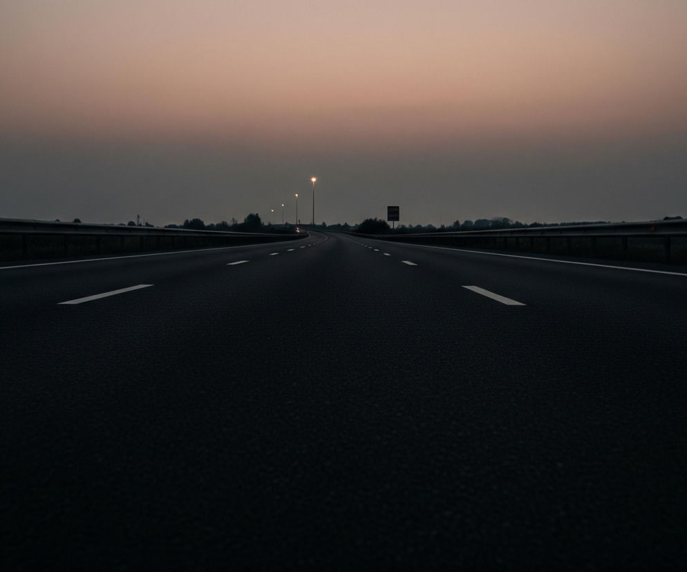 Asfaltowa autostrada rozciąga się w perspektywie, z widocznymi przerywanymi białymi liniami pasów ruchu. Po obu stronach drogi biegną metalowe barierki ochronne, za którymi widać pasy zieleni i drzewa. Horyzont dominuje rozmyte, bladopomarańczowe niebo, stopniowo przechodzące w ciemny szary, a na horyzoncie z lewej strony widoczne są pojedyncze, świecące lampy uliczne.