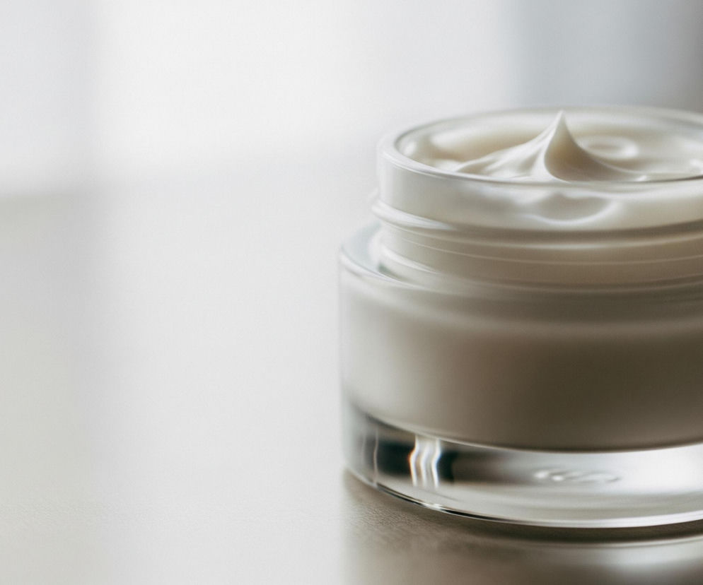 W prawym centrum obrazu znajduje się otwarty, przezroczysty szklany słoik wypełniony białą kremową substancją. Krem wystaje ponad krawędź słoika, tworząc zawiłą, stożkowatą formę na środku. Słoik ma grubą, przezroczystą podstawę i jest częściowo widoczny z prawej strony kadru, gdzie ciemniejszy element zasłania jego część. Lewa strona obrazu jest rozmyta i jasna, tworząc miękkie, białe tło. Cały obiekt spoczywa na gładkiej, lekko błyszczącej powierzchni, odbijającej światło.