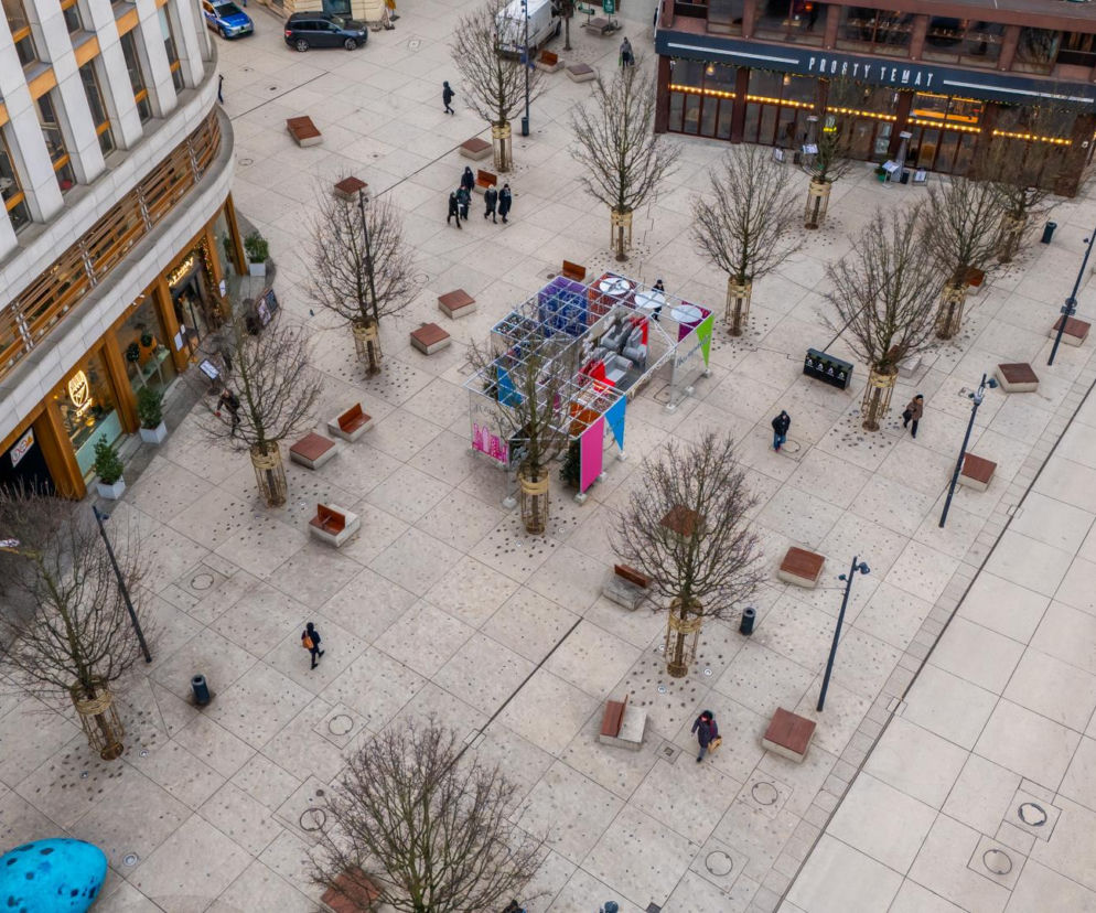 Widok z góry na Plac Pięciu Rogów w Warszawie zimą, z gołymi drzewami, betonową nawierzchnią, kilkoma przechodniami i pustymi ławkami. Widoczna jest tymczasowa, kolorowa instalacja artystyczna na środku placu. Więcej o Nowym Centrum Warszawy przeczytasz na Architektura Murator Plus.