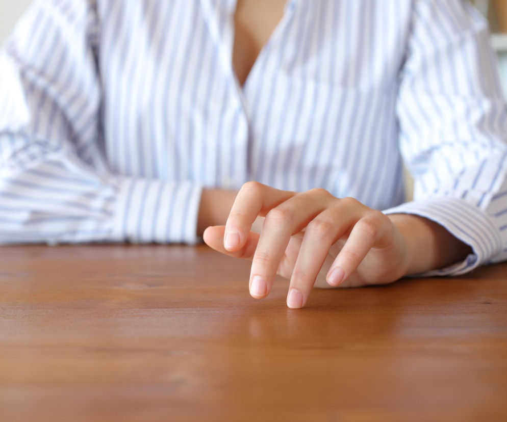 Dłoń kobiety w biało-niebieskiej koszuli, palcami delikatnie stuka o brązowy, drewniany blat biurka, co symbolizuje nieświadomy nawyk „fidgeting”, poprawiający koncentrację. Więcej o wpływie drobnych ruchów na skupienie przeczytasz na Poradnik Zdrowie.