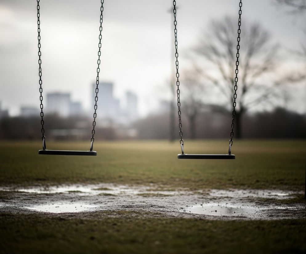 Na pierwszym planie widoczne są dwa puste siedziska huśtawek, wiszące na metalowych łańcuchach przymocowanych do konstrukcji z ciemnych słupków. Poniżej rozciąga się zielono-brązowa trawa z wyraźnymi kałużami odbijającymi światło. W tle, częściowo rozmyte, majaczą zarysy drzew bez liści i odległe, niewyraźne budynki o szarych fasadach, wszystko pod jasnym, zachmurzonym niebem.