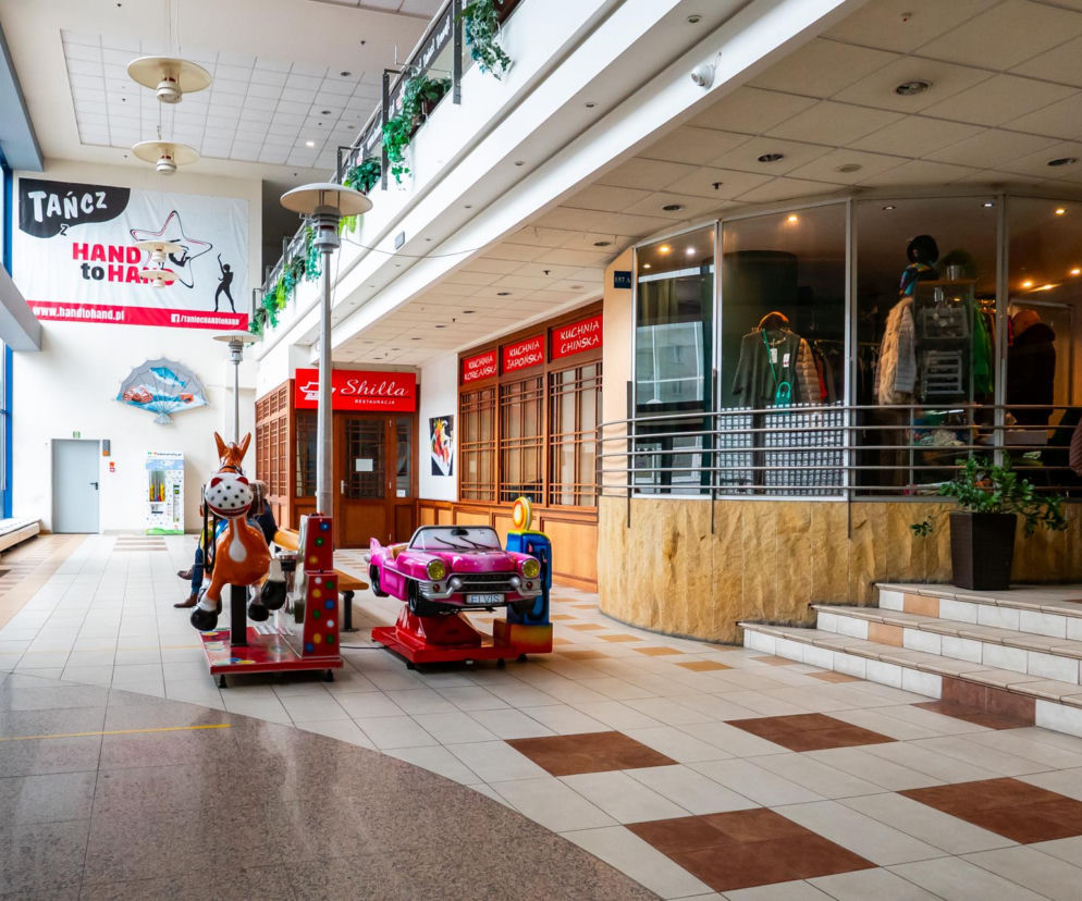 Wnętrze CH Land w Warszawie, ukazujące retro estetykę lat 90. Widoczne automaty do zabawy dla dzieci, sklep odzieżowy, restauracja Shilla oraz puste lokale. To świadectwo przemijającej architektury handlowej, o której więcej przeczytasz na Architektura Murator Plus.