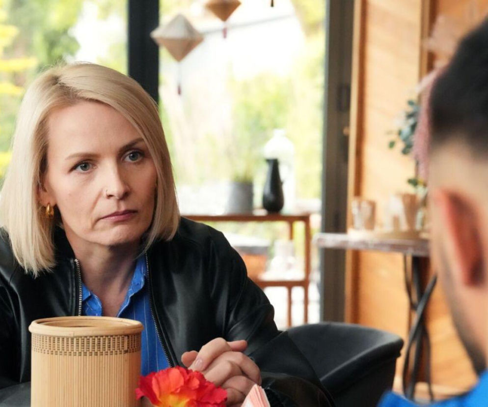 Kobieta z blond włosami, Olga (Dorota Łukasiewicz-Kwietniewska), ubrana w czarną skórzaną kurtkę i niebieską koszulę, siedzi przy stole, patrząc z powagą na mężczyznę, którego widać z tyłu głowy. Przed nią widać wiklinowy pojemnik i czerwony kwiat. Scena z serialu „Barwy szczęścia” dostępna na portalu Super Seriale.