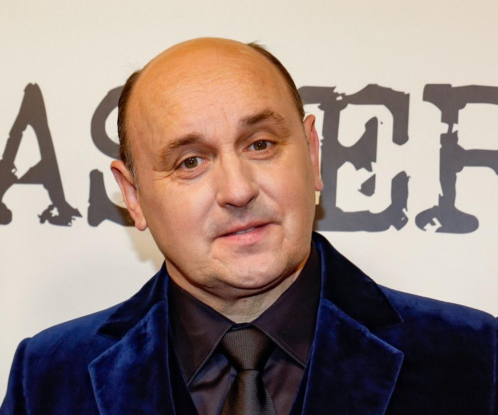 Aktor Adam Woronowicz w granatowej welurowej marynarce i ciemnej koszuli z krawatem. O programie Taskmaster przeczytasz na SE.