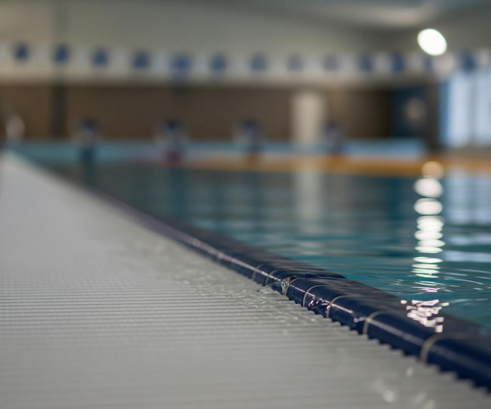 Na zdjęciu widoczna jest krawędź basenu, z błękitną wodą wypełniającą prawą stronę kadru i jasnoszarym, antypoślizgowym wykończeniem po lewej stronie. Na granicy wody i brzegu biegnie ciemnoniebieska lina torowa z falującymi elementami. Woda delikatnie faluje, odbijając rozmazane, jasne światła z sufitu. W tle, poza basenem, widoczne są rozmyte, niebiesko-białe znaczniki torów, a dalej szklane ściany i czerwonawa boja ratunkowa.