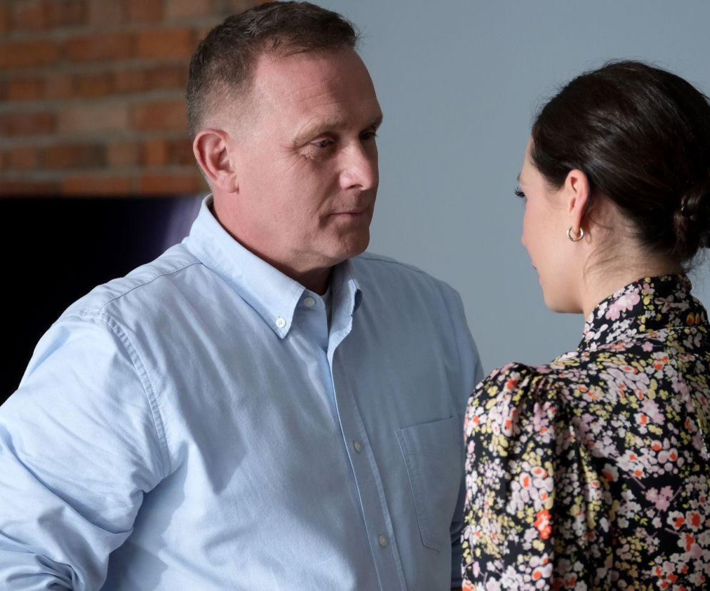 Artur Rogowski (Robert Moskwa) w błękitnej koszuli patrzy na Joannę Dobrzańską (Dominika Sakowicz) w kwiecistej bluzce z upiętymi włosami. Para rozmawia w tle ceglanej ściany. Scena ilustruje konflikt Rogowskiego między rodziną a nowym romansem, o którym czytaj na Super Seriale.