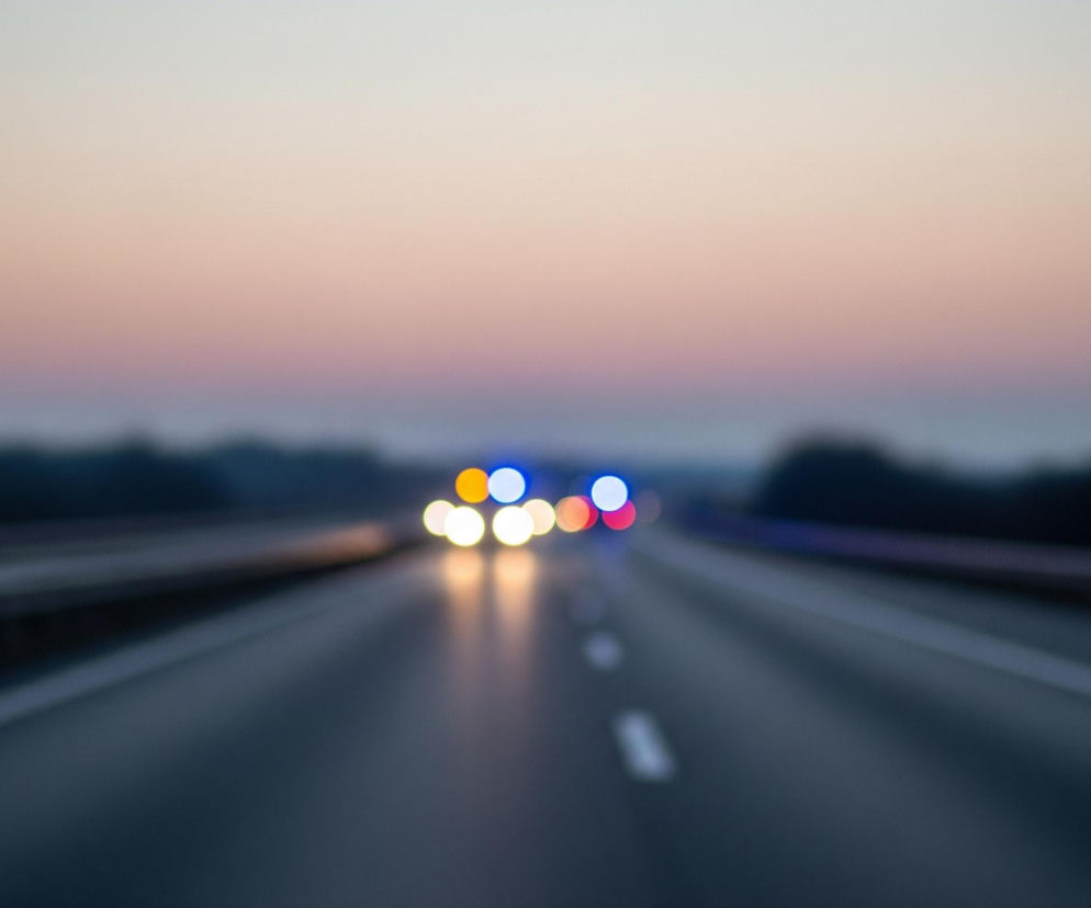 Autostrada rozciąga się w głąb obrazu, a jej asfaltowa nawierzchnia jest ciemnoszara, z rozmytymi białymi liniami przerywanymi pośrodku. Po obu stronach drogi widoczne są niewyraźne, ciemne bariery ochronne, za którymi rozciąga się rozmyty pas ciemnych drzew lub krzewów. W oddali, na horyzoncie, znajdują się rozmyte światła pojazdów: białe i żółte reflektory, a także niebieskie i czerwone światła sygnalizacyjne. Nad autostradą rozciąga się niebo o delikatnych, pastelowych odcieniach, przechodzących od jasnego różu i pomarańczu przy horyzoncie do jaśniejszej, niemal białej barwy w górnej części kadru.