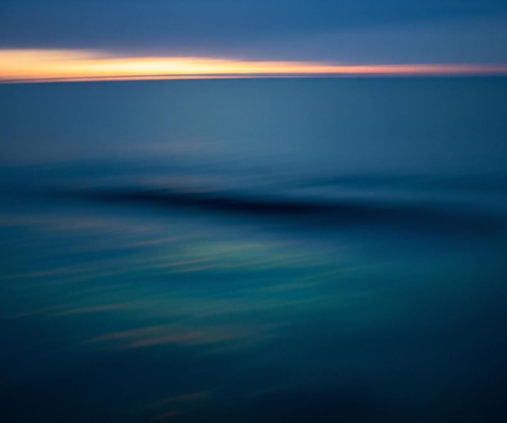 Dominujący na obrazie jest kolor niebieski w różnych odcieniach, od ciemnego granatu na dole, po jaśniejsze, turkusowe partie w środkowej części, aż do głębokiego błękitu u góry. Horyzont przecina pozioma, rozmyta smuga światła w odcieniach pomarańczu i żółci, stanowiąca wyraźny, cieplejszy akcent. Poniżej horyzontu widoczne są rozmazane, podłużne kształty w ciemniejszym odcieniu niebieskiego, sugerujące ruch wody lub mgły. Cały obraz ma miękkie, rozmyte krawędzie i jest utrzymany w nastrojowej, spokojnej tonacji.