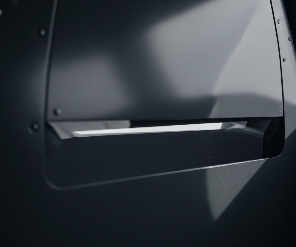 Ciemnoszare, matowe płaszczyzny z metalowymi nitami widoczne są na całym obrazie, tworząc industrialny, ascetyczny kontekst. Centralną część zajmuje prostokątne wycięcie z zaokrąglonymi rogami, które odsłania jaśniejszy, poziomy pasek, sugerujący otwartą szczelinę lub okno. Na górnej i prawej krawędzi wycięcia oraz na lewej pionowej płaszczyźnie rozmieszczone są rzędy błyszczących, srebrnych nitów. Światło padające z prawego górnego rogu rozjaśnia częściowo wnętrze wycięcia i tworzy subtelne refleksy na powierzchniach.