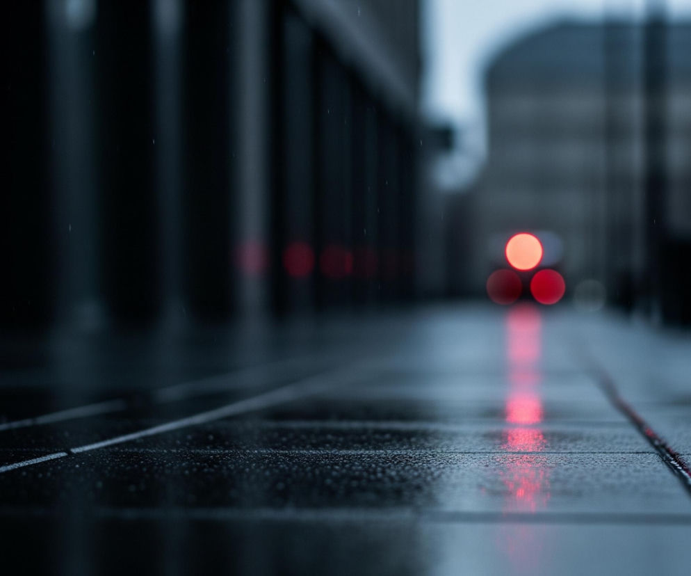 Chodnik z szarych, prostokątnych płyt jest mokry i pokryty drobnymi kroplami wody, odbijającymi światło. Na nim widoczne są rozmyte, intensywnie czerwone refleksy świetlne, ułożone w linię, która biegnie od środka kadru w głąb. W tle, na końcu chodnika, znajdują się niewyraźne, okrągłe, czerwone światła, przypominające lampy uliczne lub światła pojazdu. Po lewej stronie widać ciemne, rozmazane pionowe kształty, sugerujące budynki lub słupy. Całość utrzymana jest w chłodnych, ciemnych odcieniach szarości i błękitu, z kontrastującymi czerwonymi akcentami.