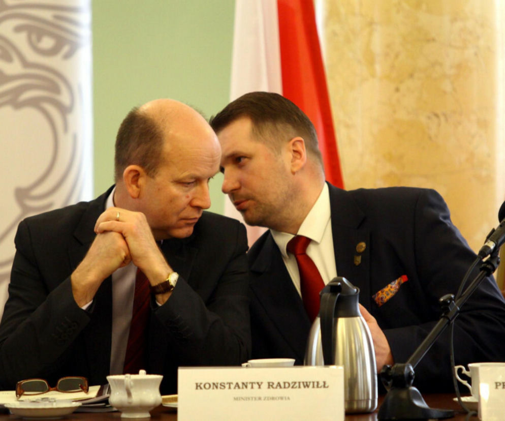 Przemysław Czarnek i Konstanty Radziwiłł rozmawiają podczas spotkania. Czarnek, w garniturze i krawacie, pochyla się w stronę Radziwiłła, który siedzi z dłońmi splecionymi na stole. Na stole widać mikrofon, karafkę oraz kartkę z napisem Konstanty Radziwiłł Minister Zdrowia. Więcej informacji o majątku Czarnka znajdziesz na Super Biznes.