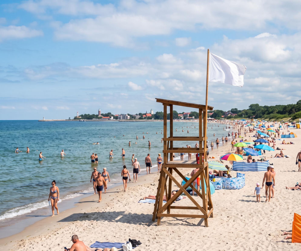 Wieża ratownicza z białą flagą na pełnej ludzi plaży w Darłowie. O otwartych kąpieliskach dowiesz się z SE Szczecin.