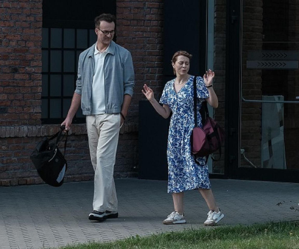 Aktor Łukasz Wójcik jako Radomir i Karina Seweryn jako Dorota z serialu Barwy szczęścia idą razem. Mężczyzna w jasnych spodniach i kurtce, kobieta w niebieskiej sukience w białe wzory, z czerwoną torbą. Scena z odcinka, o którym przeczytasz więcej na Super Seriale.