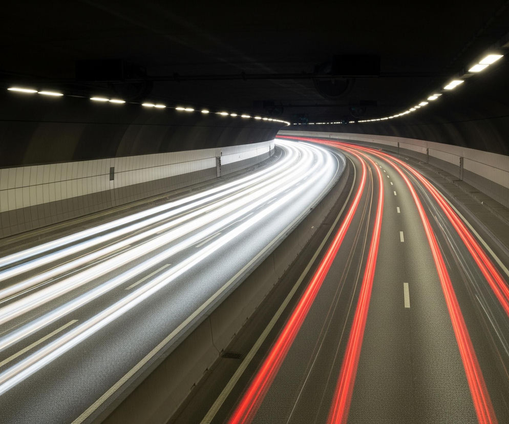 Wewnątrz ciemnego tunelu autostradowego, oświetlonego rzędami jasnych lamp wzdłuż zakrzywionego sufitu, widoczne są dwa pasy ruchu rozdzielone białą linią przerywaną. Po lewej stronie intensywne, białe smugi światła, biegnące prosto, świadczą o ruchu pojazdów w kierunku obserwatora. Po prawej, czerwone smugi światła, delikatnie zakrzywione, wskazują na ruch pojazdów oddalających się w głąb tunelu. Ściany tunelu, obłożone jasno-szarymi panelami u dołu, przechodzą w ciemniejsze odcienie ku górze, gdzie styka się z czarnym sufitem.