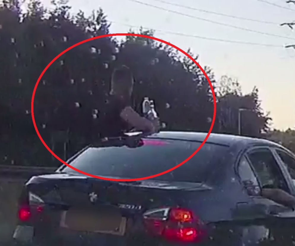 Jeden z pasażerów samochodu pije alkohol opierając się o dach pojazdu