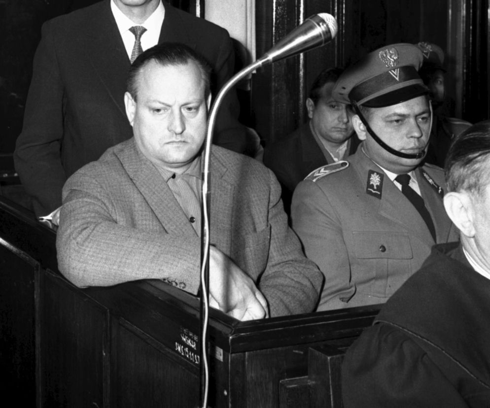 Stanisław Wawrzecki, główny oskarżony w aferze mięsnej, siedzący na ławie oskarżonych podczas procesu sądowego. Jego twarz wyraża powagę i skupienie. U jego boku, po prawej stronie, widać mężczyznę w mundurze milicjanta. Więcej o aferze przeczytasz na naszym portalu.