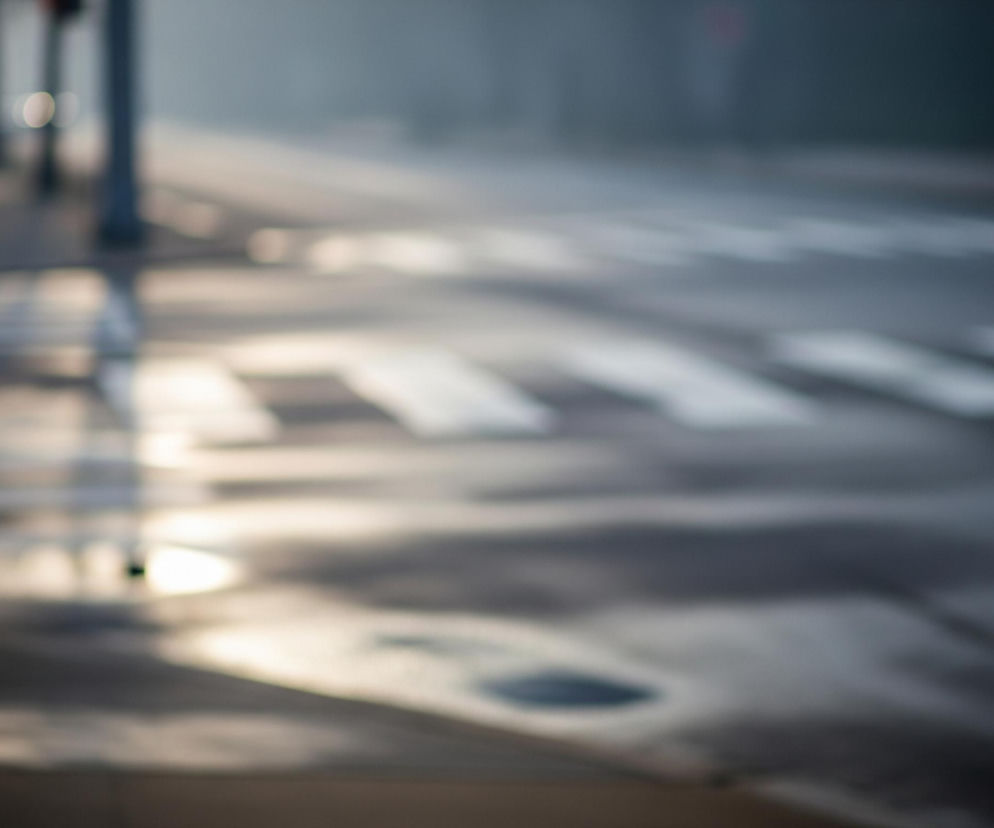 Droga jest mokra, pokryta plamami światła i kałużami, z których jedna widoczna jest w prawym dolnym rogu. Na czarnej, rozmytej jezdni widać białe pasy przejścia dla pieszych, ułożone równolegle. Po lewej stronie widać niewyraźny słupek lub słup. Całość obrazu jest rozmyta, utrzymana w ciemnych, szarych i beżowych tonach, z wyraźnymi jasnymi odbiciami na mokrej powierzchni.