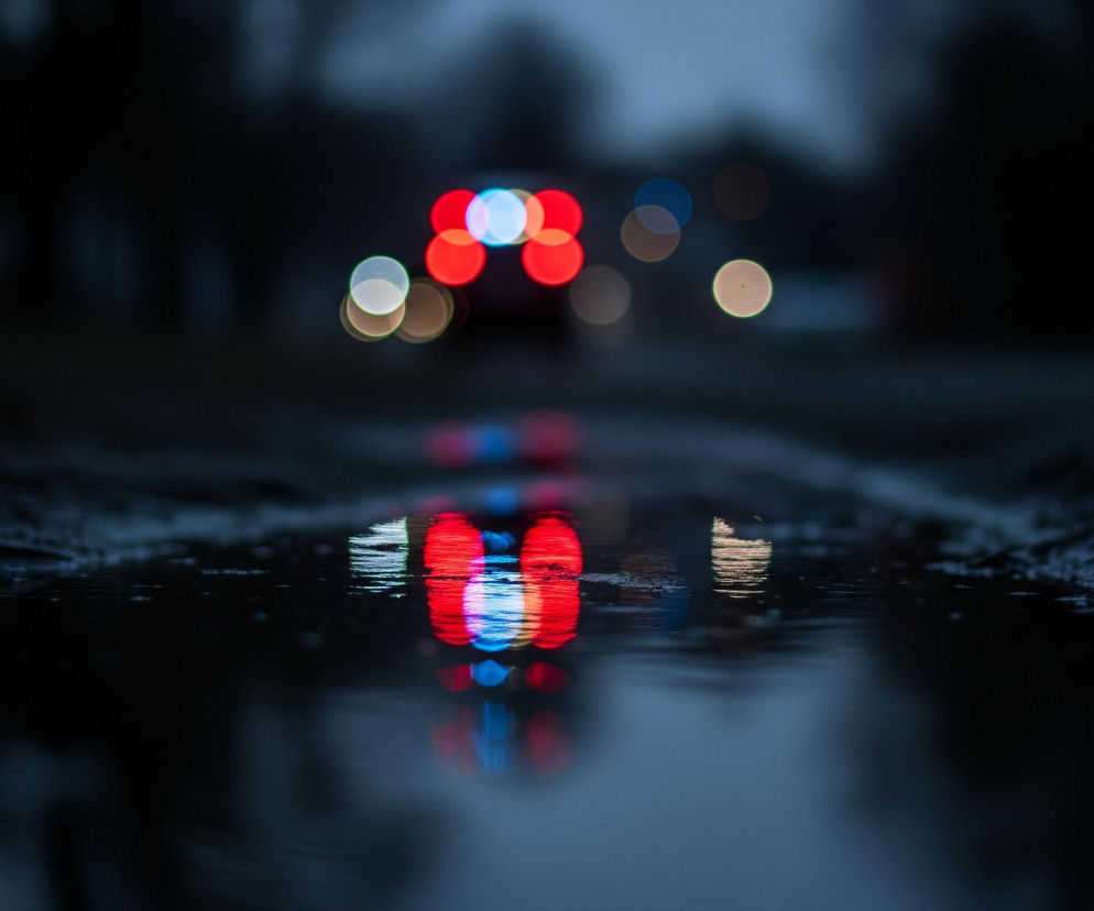 Głęboka kałuża zajmuje dużą część pierwszego planu, odbijając rozmazane światła znajdujące się w oddali. Na ciemnym, rozmytym tle widoczne są okrągłe, kolorowe refleksy świetlne: dominują czerwone i niebieskie, ale pojawiają się też pomarańczowe i białe. Te barwne kręgi rozmywają się w pionowe smugi na powierzchni wody, tworząc efektowne odbicie świetlnych punktów. Powierzchnia drogi wokół kałuży jest nierówna i matowa, o ciemnej, brudnej teksturze.